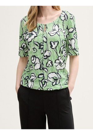 Green floral blouse featuring a loose fit, round neckline, and keyhole detail with dark outlines on a textured fabric. Paired with black pants.