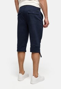 Navy blue knee-length shorts made of soft fabric. Features rolled cuffs, two back pockets, and subtle hardware accents. Clean design.