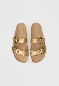 Χρυσά ψεύτικα δερμάτινα σανδάλια με επίπεδη σόλα, δύο ρυθμιζόμενες αγκράφες στις σχέσεις και κρεμ σουέτ πέλμα.