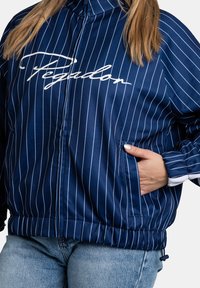 Granatowa kurtka zapinana na zamek z białymi pionowymi paskami, posiadająca wysoki kołnierz, ściągacze na mankietach oraz logo na piersi. Zawiera kieszenie po bokach.