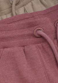 Deux paires de pantalons de survêtement empilées, l'une en rose doux et l'autre en beige. Dotées de ceintures côtelées avec un cordon de serrage, mettant en valeur des textures mélangées.