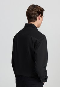 Czarna, długosleeve'owa koszula z kołnierzykiem, o gładkiej teksturze i skróconym fasonie, widziana z tyłu. Proste mankiety zapinane na guziki.