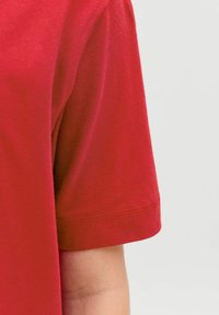 Közelről egy személy karja, aki rövid ujjú piros inget visel, világosszürke háttér előtt.