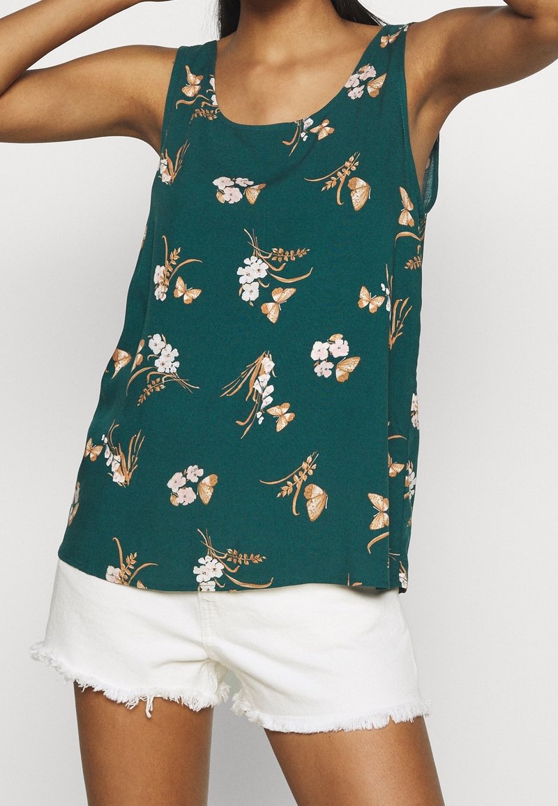 Haut sans manches bleu sarcelle avec imprimé floral blanc et marron et papillons, porté avec un short blanc effiloché, les bras du modèle levés au-dessus des épaules.