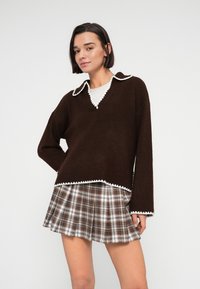 Καφέ πλεκτό πουλόβερ με λευκή γιακαδάκι με κυματιστούς κυματισμούς, συνδυασμένο με πλισέ καρό φούστα σε καφέ, λευκούς και γκρι τόνους.