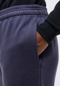 Σκούρο μπλε φόρμα με ελαστική μέση και πλαϊνή τσέπη που κρατά το χέρι ενός ατόμου που φοράει μαύρο μακρυμάνικο μπλουζάκι, με το κέντημα "BORG" στο πόδι.