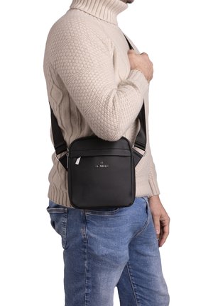 Borsa a tracolla nera con texture liscia e chiusura con zip, indossata a spalla; dotata di tracolla regolabile e logo frontale.