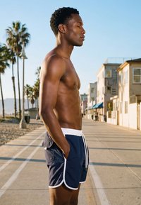 Muž bez košile se zavřenýma očima a rukama v kapsách stojí na sluncem zalité stezce u pláže lemované palmami a budovami.