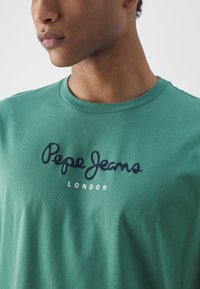 Zöld pamut póló kerek nyakkivágással, amelyen a "Pepe Jeans" felirat navy színben, alatta pedig a "LONDON" felirat fehér színben található. Sima textúra.