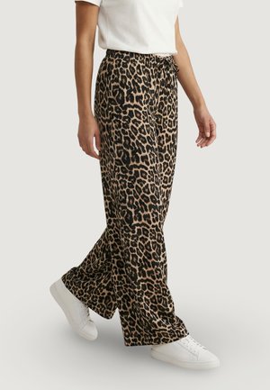 Persona che indossa pantaloni a gamba larga con stampa leopardata, scarpe da ginnastica bianche e una maglietta bianca a maniche corte infilata, cammina davanti a uno sfondo semplice.