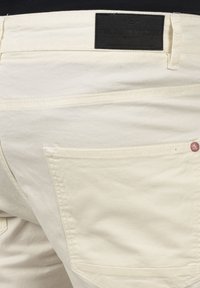 Lyse beige bukser laget av bomull, med en teksturert finish, baklommer og et svart lærmerke i midjen med merkeinformasjon.