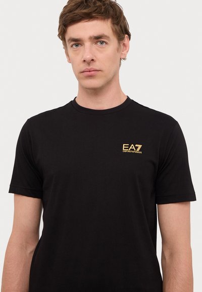 T-shirt noir à manches courtes en coton doux, avec un logo EA7 doré sur la poitrine. Col rond classique, texture lisse, design minimaliste.