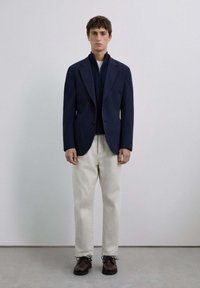 Blazer azul marino sobre una chaqueta oscura con cremallera, pantalones crema y zapatos marrones con cordones; diseño minimalista con texturas suaves y corte ajustado.