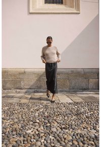 Pull en ribbed beige clair, jupe maxi en satin noir, ballerines noires avec des accents, lunettes de soleil, marchant sur un chemin de galets près d'un mur clair.