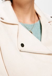 Jasny beżowy żakiet z bliskim ujęciem suwaka, srebrnymi detalami i czarnymi zatrzaskami, nałożony na fakturowaną jasno-niebieską bluzkę.