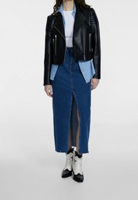 Giacca da biker nera in pelle con dettagli in cerniera, camicia di jeans azzurri sotto, e lunga gonna di jeans blu con spacco frontale. Stivali bianchi e neri.