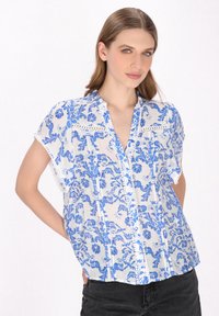 Let og kortærmet bluse med et hvidt stof med blå blomster mønstre, en klipdecollar og delikat blonde detalje på skødet.