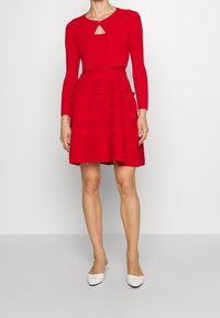 Robe rouge côtelée avec jupe à volants superposés et cardigan court avec un détail en trou de serrure. Associée à des chaussures slingback blanches.