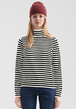Junge Frau mit roter Strickmütze, klaren Brillen, schwarz-weiß gestreiftem Langarmshirt und dunkelblauen Jeans steht vor einem schlichten Hintergrund.