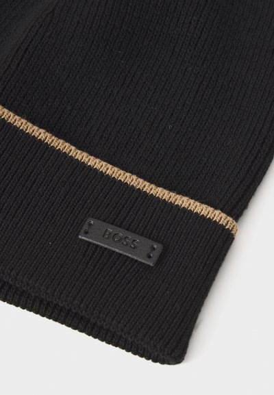 Bonnet en tricot noir avec une surface texturée, arborant une rayure dorée contrastante et une étiquette logo en relief noire. Fabriqué en matériau doux et flexible.