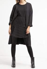 Cardigan en tricot noir sur un haut noir, associé à une jupe noire à la forme unique avec des rayures horizontales et un collant noir, le tout accompagné de bottines.