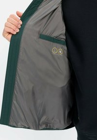 Vandafvisende jakke med en grøn yderskal og en grå inderside. Har en lomme og ikoner, der angiver vand- og pletmodstand.