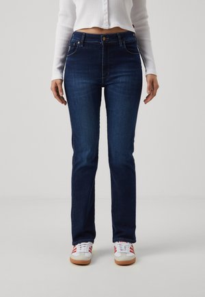 Persoon die donkerblauwe jeans met rechte pijpen, een witte geribbelde cropped blouse met knopen en witte sneakers met rode strepen draagt, staand tegen een effen achtergrond.