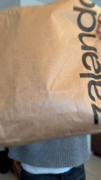 Brunt papirindpakning med sort tekst "zalando", med folder og krøller, holdt af en person iført en grå trøje.