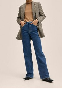 Blazer a cuadros sobre un suéter de cuello alto marrón, combinado con jeans de tiro alto y pierna ancha azul, y botines negros de punta afilada.