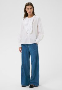 Vrouw in een witte rimpelige blouse met lange mouwen, brede blauwe jeans en zwarte ballerina's, staand tegen een egale achtergrond.