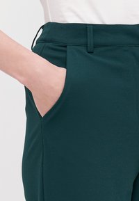 Tmavozelené nohavice s hladkou textúrou, s bočnými vreckami a skladaným dizajnom v páse. Čisté línie s minimálnymi detailami.
