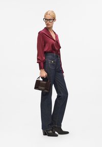 Bordeauxfarbene Satin-Bluse mit Schleifendetail, hochtaillierte dunkle Jeans, schwarze Knöchelstiefel und eine kleine dunkle Handtasche mit goldenen Akzenten.