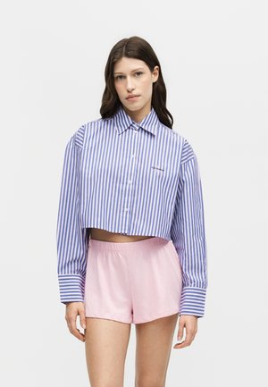 Joven mujer con una camisa corta de botones a rayas verticales azules y blancas y pantalones cortos rosa claro sobre un fondo liso.