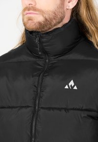 Svart vadderad jacka med hög krage, dragkedjestängning och logotypdetalj. Slät yta och quiltat mönster synligt på tyget.