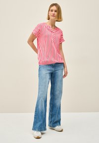 Rød og hvid stribet kortærmet bluse med klapkrave, parret med lyseblå flagrende jeans og hvide sneakers på en ensfarvet baggrund.