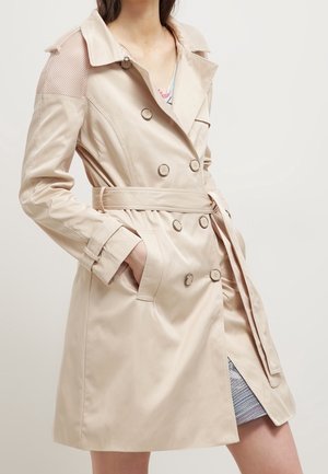 Trench-coat beige à double boutonnage avec ceinture, détails de boutons, panneaux en maille aux épaules et poches latérales, porté sur une robe à motifs.