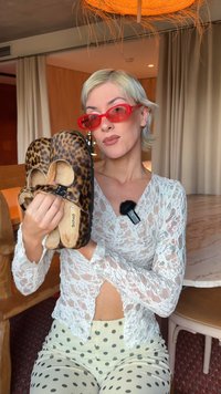 Žena s krátkými blond vlasy a červenými slunečními brýlemi drží pantofle s leopardím potiskem v místnosti s dřevěným nábytkem a mramorovým stolem.