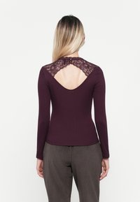 Langarmtop in donker bordeaux ribfluweel met kantdetails op de rug, voorzien van een ronde uitsparing en knoopsluiting bij de hals.