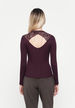 Haut à manches longues en tissu côtelé bordeaux foncé avec des détails en dentelle au dos, mettant en avant une découpe arrondie et une fermeture à boutons au cou.