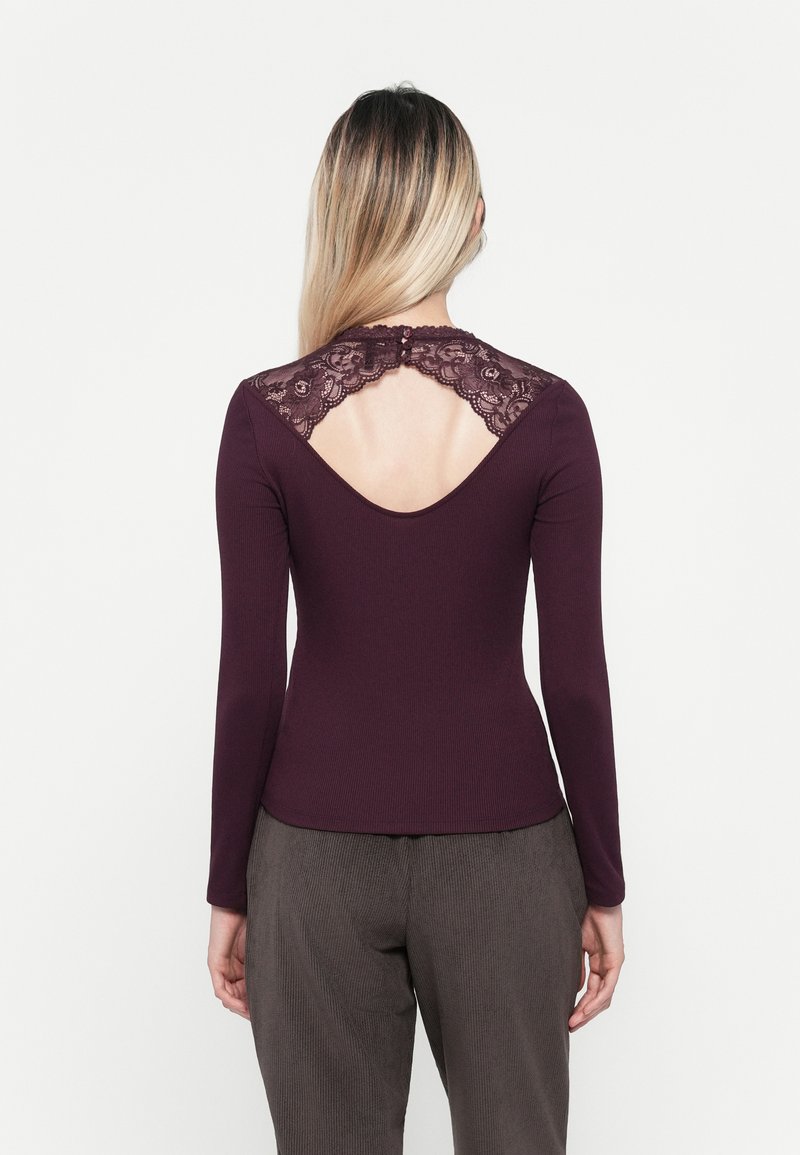 Langarmtop in donker bordeaux ribfluweel met kantdetails op de rug, voorzien van een ronde uitsparing en knoopsluiting bij de hals.