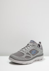 Gray athletic shoe with mesh upper, patterned accents, and a white rubber sole. Features blue logo detail and lace-up front.