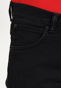 Černé džínové kalhoty s klasickým designem pěti kapes, mírně texturované s kovovými ozdobami. Pas sedí nad červenou látkou.