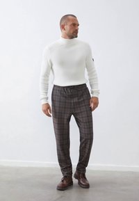 Weißer gerippter Strickpullover mit Stehkragen, kombiniert mit braunen Karohosen und braunen Schnürschuhen. Glatte Textur mit schwarzer elastischer Taillenband-Details.