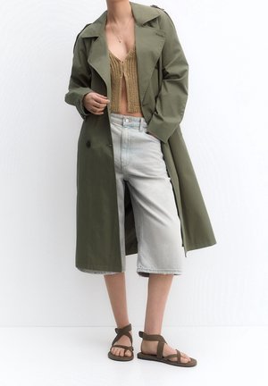 Person wearing olive green trench coat, tan knitted cropped top, light denim knee-length shorts, and brown strappy sandals standing against white background.