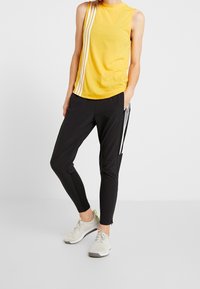 Top sin mangas amarillo con rayas laterales blancas, combinado con joggers negros con rayas laterales blancas y zapatillas deportivas elegantes con detalles en gris.