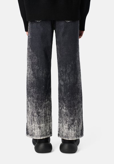 Weite Jeans aus Denim mit einem angeschnittenen Schwarz-Weiß-Gradient. Roh geschnittene Säume und eine strukturierte Oberfläche. Getragen mit schwarzen Schuhen.