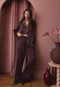 Satynowy burgundowy zestaw piżamowy z białym wykończeniem, składający się z koszuli na guziki oraz spodni z troczkami. Gładka tekstura, luźny krój, zestawiony z nowoczesnym wystrojem.