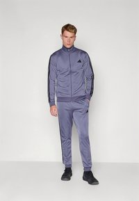 Fato de treino cinza feito de material liso; casaco com fecho de correr e riscas pretas, calças a condizer com riscas laterais e detalhes em preto, combinado com sapatos pretos.