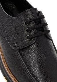 Chaussure en cuir noir avec une surface texturée, dotée de deux œillets en métal, de lacets noirs et d'un détail de couture contrastante en marron le long de la semelle.