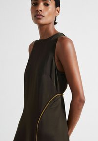 Ärmelloses dunkelbraunes Kleid mit einer glatten Textur, schrägen Nähten und einem kontrastierenden goldenen Reißverschlussdetails an der Seite.